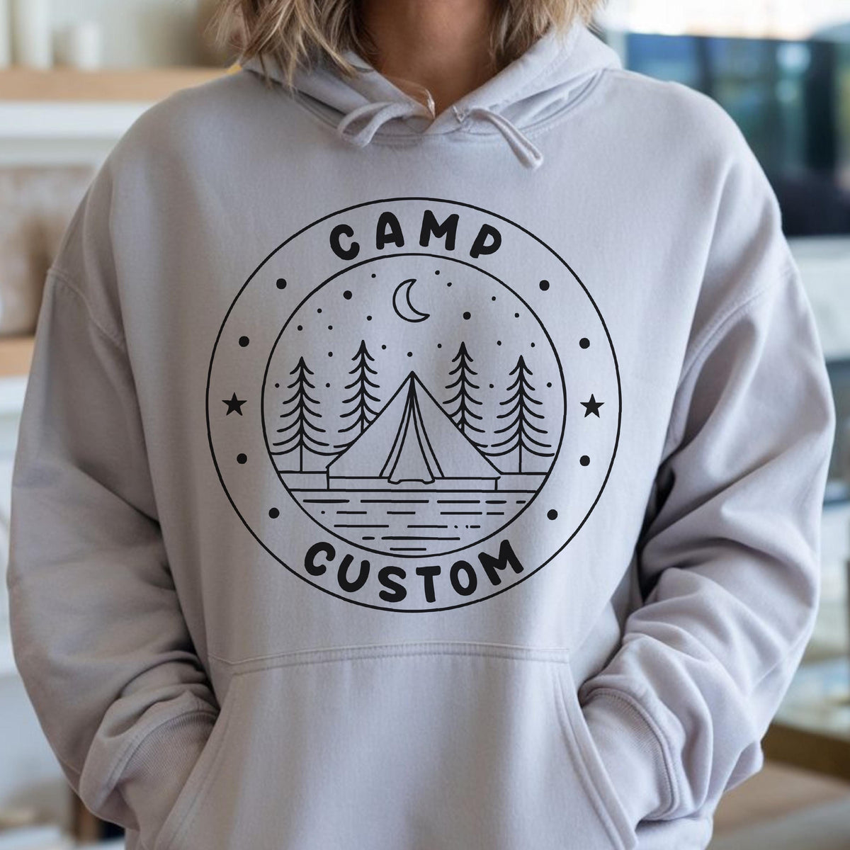 May include: A light gray hooded sweatshirt featuring a black circular graphic on the chest. The design includes a tent, pine trees, a crescent moon, and stars, with the text CAMP at the top and CUSTOM at the bottom of the circular border.