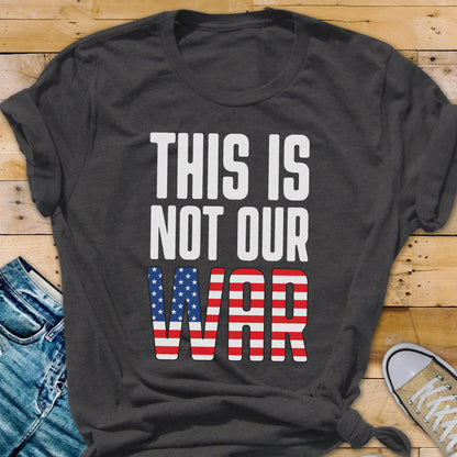 May include: Dark gray t-shirt with the text "THIS IS NOT OUR WAR" in white. The word "WAR" is designed with the American flag's stars and stripes. The shirt is folded at the sleeves and knotted at the bottom.