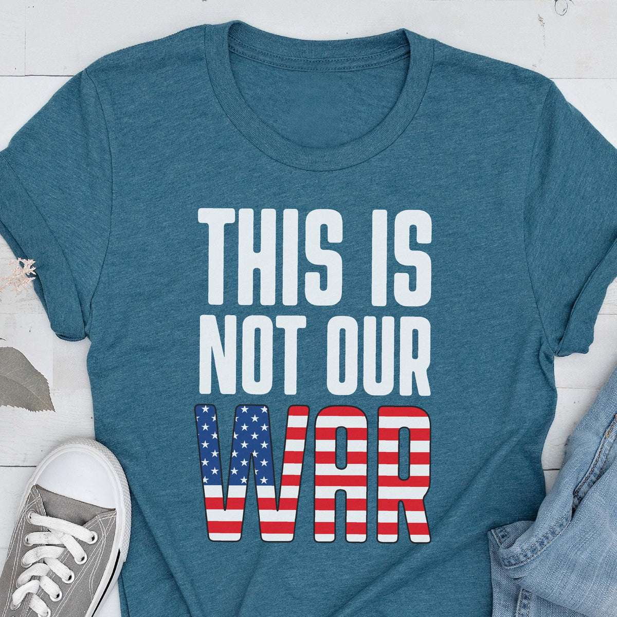 May include: Teal t-shirt with the text "THIS IS NOT OUR WAR" in white block letters. The word "WAR" features a design of red and white stripes and blue stars, resembling the American flag. A gray sneaker is visible in the lower left corner.