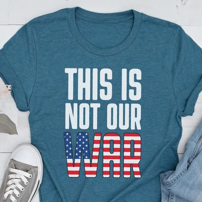May include: Teal t-shirt with the text "THIS IS NOT OUR WAR" in white block letters. The word "WAR" features a design of red and white stripes and blue stars, resembling the American flag. A gray sneaker is visible in the lower left corner.