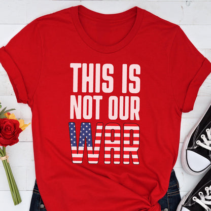 May include: A red t-shirt with the text "THIS IS NOT OUR WAR" in white block letters. The word "WAR" features a design of the American flag. The shirt is paired with blue jeans and black sneakers.