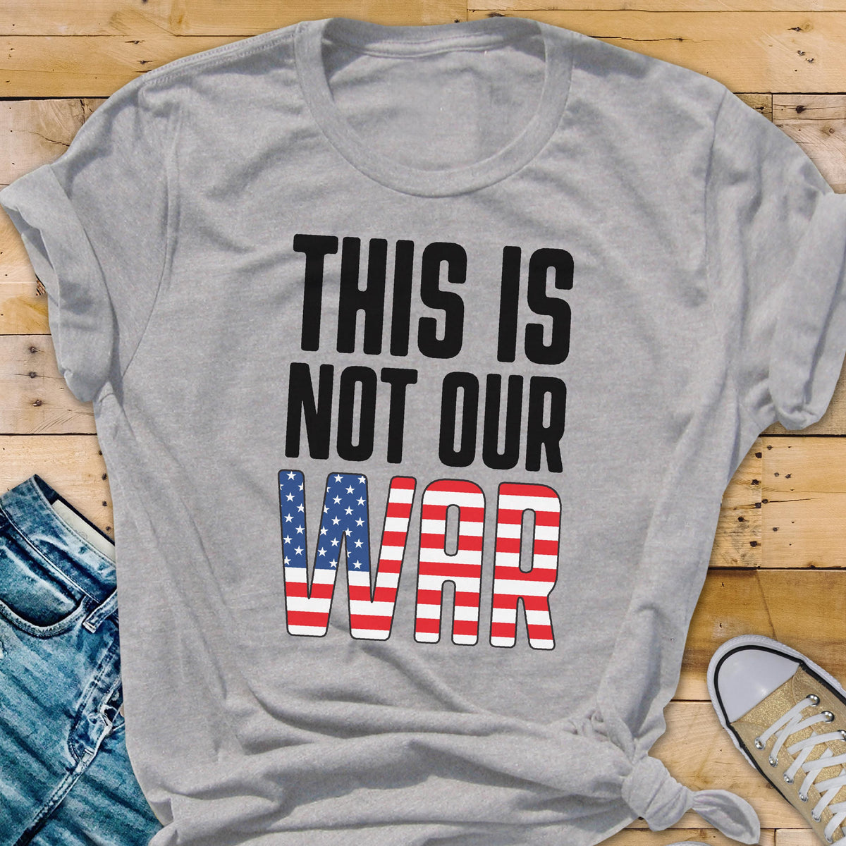May include: A gray t-shirt with the text "THIS IS NOT OUR WAR" in black block letters. The word "WAR" is designed with a red, white, and blue American flag pattern. The shirt is folded and lies on a wooden surface.