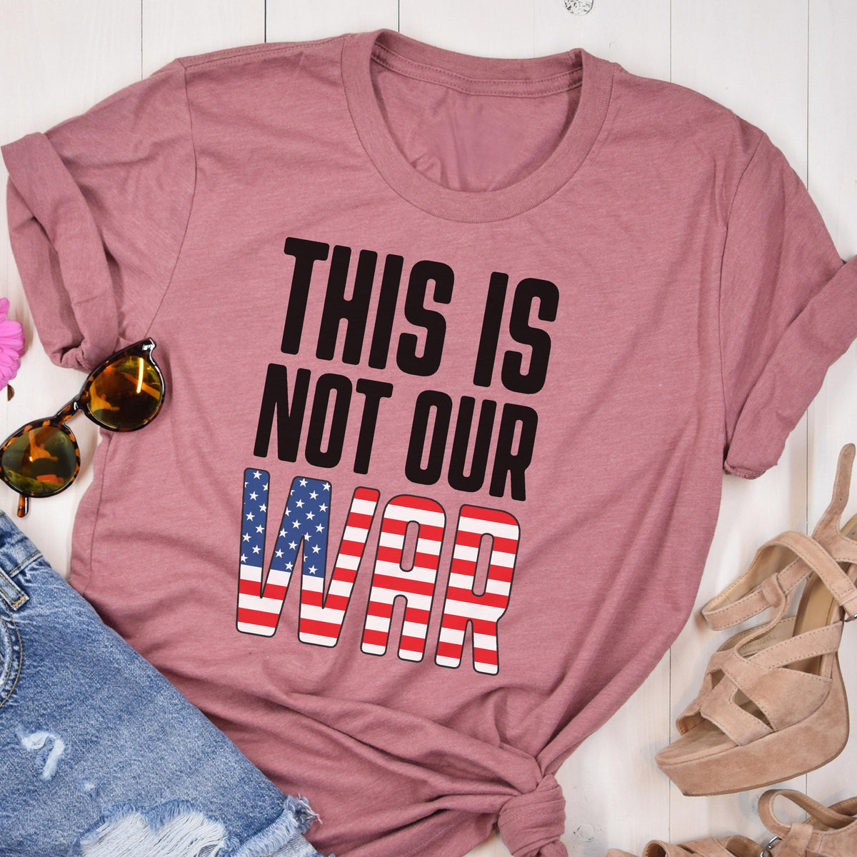 May include: A dusty rose t-shirt with the text "THIS IS NOT OUR WAR" in black and red, white, and blue. The word "WAR" is designed with the American flag. Sunglasses and denim shorts are visible.