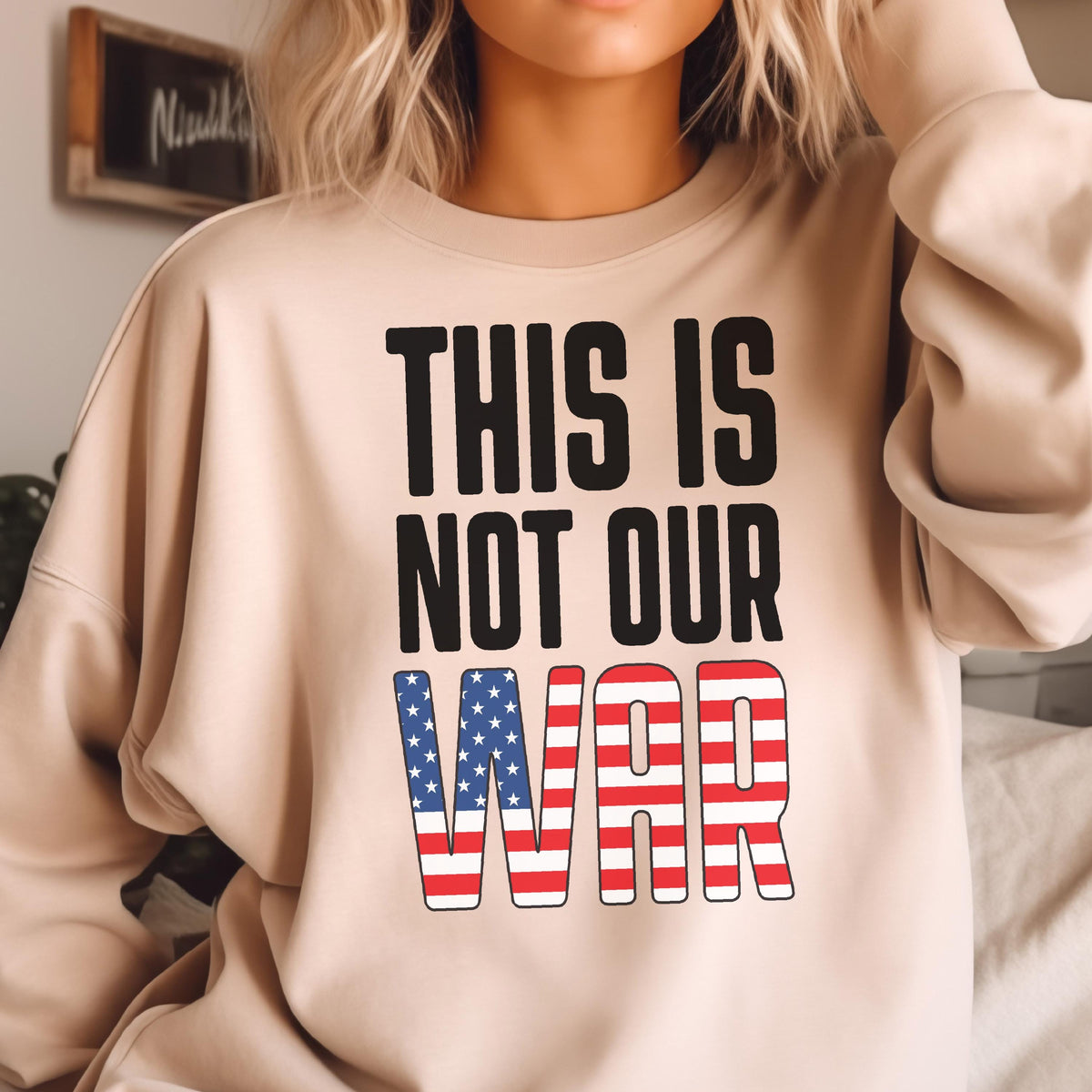 May include: Beige sweatshirt with the text "THIS IS NOT OUR WAR" in black block letters. The word "WAR" is designed with a red and white striped pattern and blue stars, resembling the American flag.
