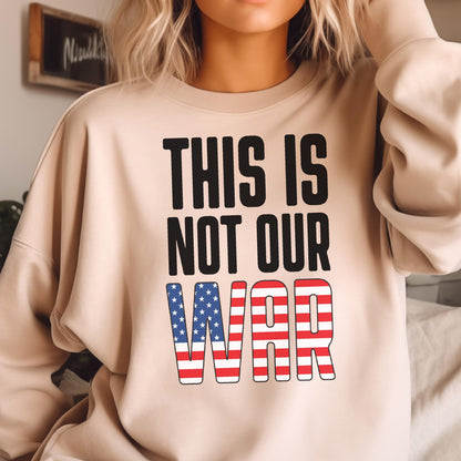 May include: Beige sweatshirt with the text "THIS IS NOT OUR WAR" in black block letters. The word "WAR" is designed with a red and white striped pattern and blue stars, resembling the American flag.