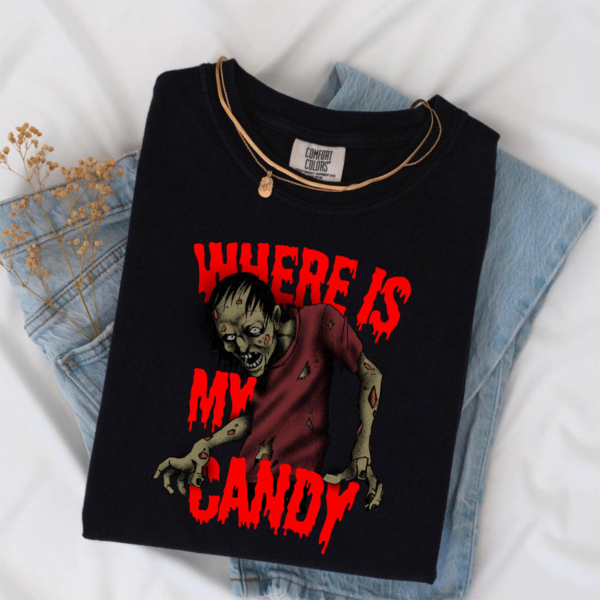 May include: Black t-shirt with a zombie graphic and the text "WHERE IS MY CANDY" in red. The zombie is wearing a red shirt. A gold necklace is draped over the shirt. The shirt is folded on top of blue jeans.