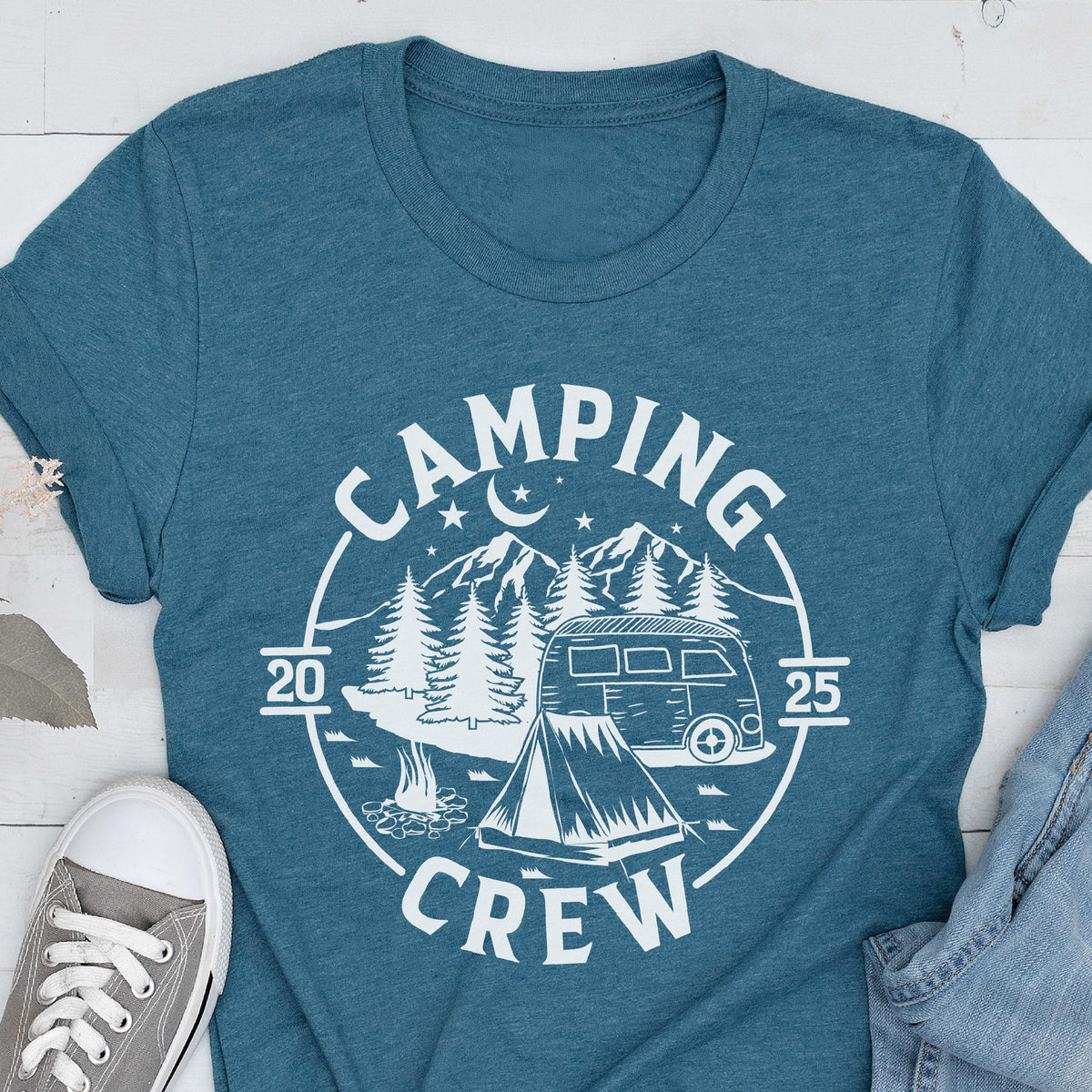 May include: A teal blue short-sleeve t-shirt featuring a white circular graphic print. The design includes a camper van, a tent, a campfire, pine trees, mountains, stars, and a crescent moon, with the text "CAMPING CREW" and "2025".