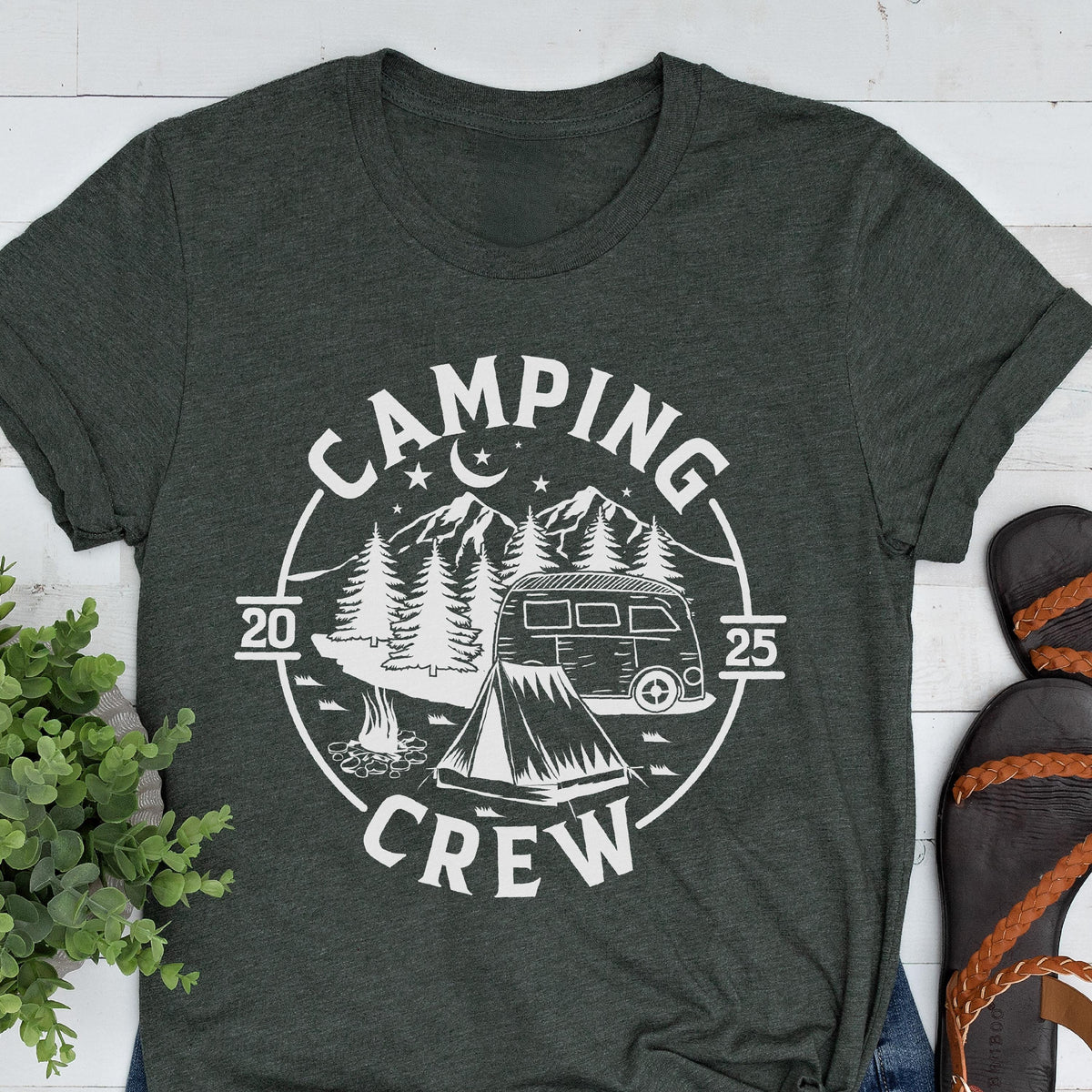 May include: A dark charcoal gray t-shirt featuring a white circular graphic with the text "CAMPING CREW" and "20 25". The design includes a camper van, a tent, pine trees, a crescent moon, and stars, all rendered in a clean, white line art style.