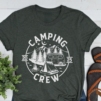 May include: A dark charcoal gray t-shirt featuring a white circular graphic with the text "CAMPING CREW" and "20 25". The design includes a camper van, a tent, pine trees, a crescent moon, and stars, all rendered in a clean, white line art style.