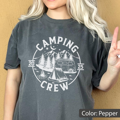 May include: A charcoal gray short-sleeve t-shirt featuring a white circular graphic with the text "CAMPING CREW" and "2025". The design includes a camper van, a tent, pine trees, mountains, a crescent moon, and stars, all printed on the front.
