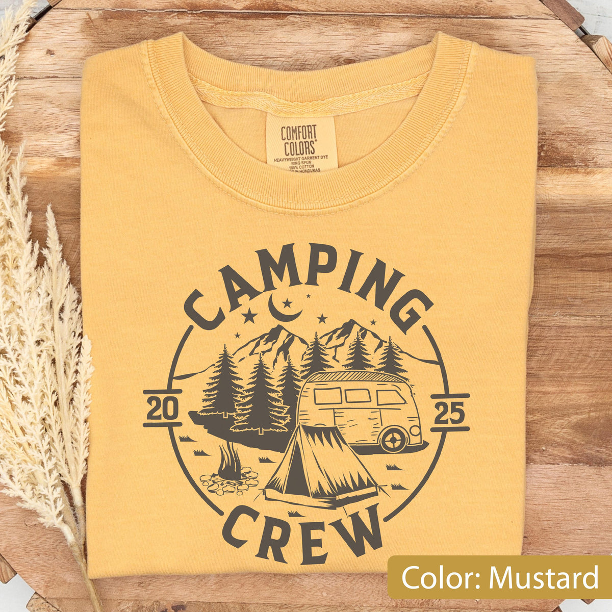 May include: A mustard yellow crew neck t-shirt featuring a dark gray circular graphic. The design includes a camper van, a tent, a campfire, pine trees, mountains, and a crescent moon with stars. The text reads CAMPING CREW 2025.