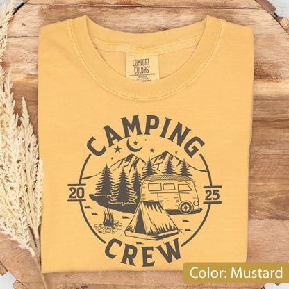 May include: A mustard yellow crew neck t-shirt featuring a dark gray circular graphic. The design includes a camper van, a tent, a campfire, pine trees, mountains, and a crescent moon with stars. The text reads CAMPING CREW 2025.