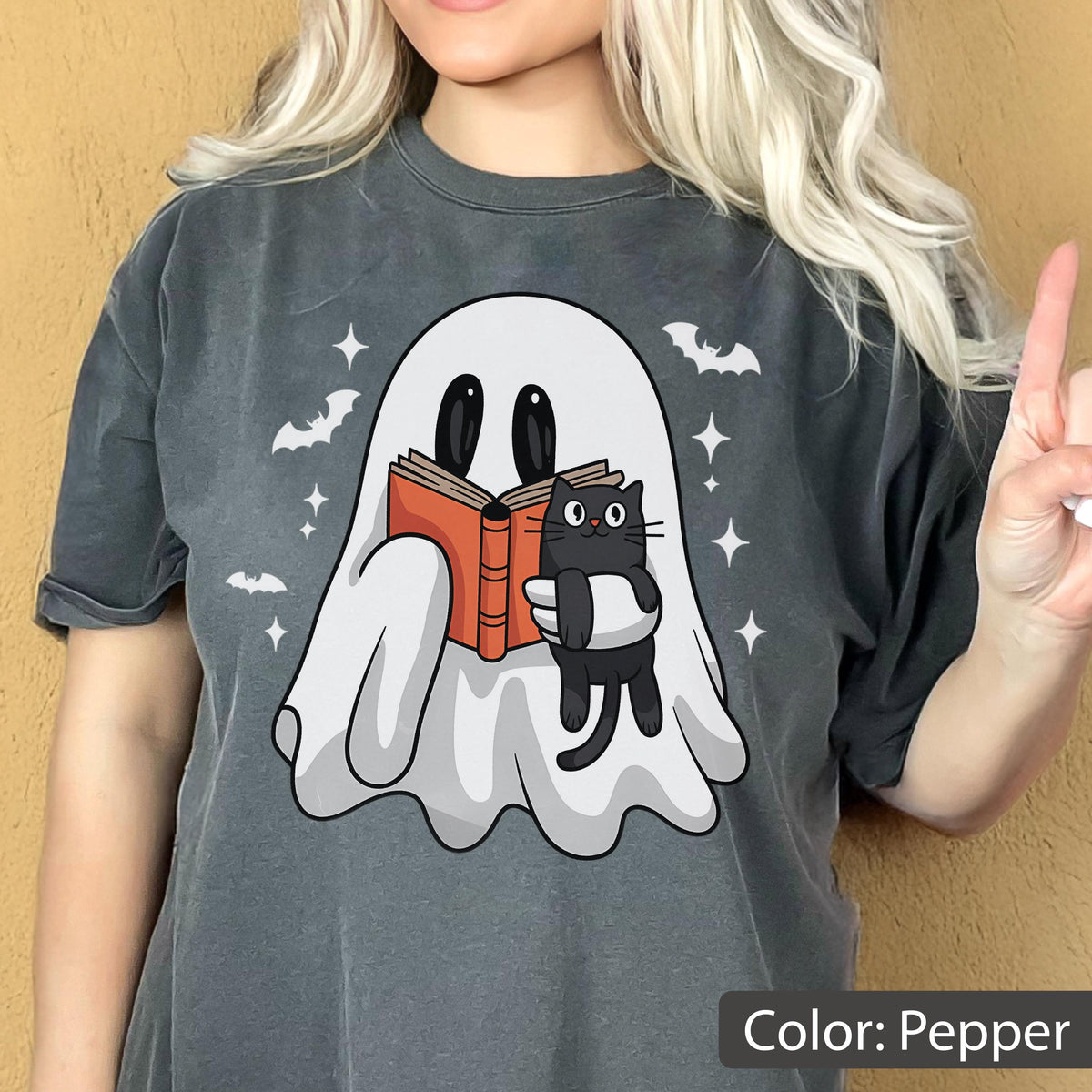 May include: Pepper-colored t-shirt featuring a cartoon ghost reading a book while holding a black cat. The design includes bats and stars. The text "Color: Pepper" is at the bottom.