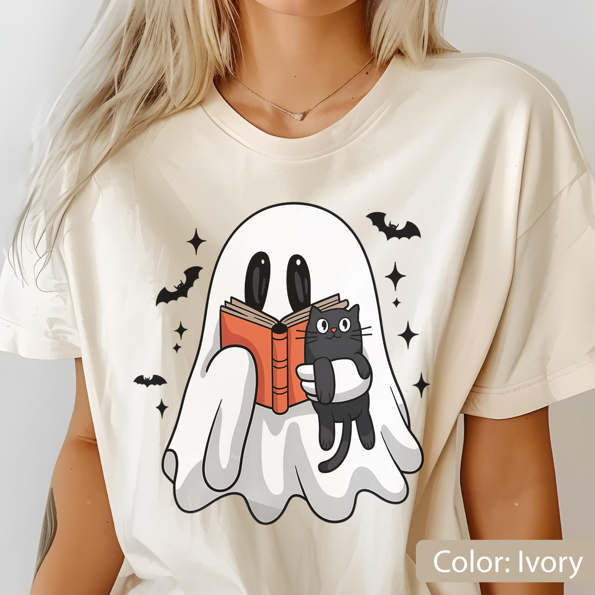 May include: Ivory-colored t-shirt featuring a cartoon ghost reading a book, with a black cat and bats. The ghost has large black eyes and is holding an open book with orange pages. Black bats and stars surround the ghost.
