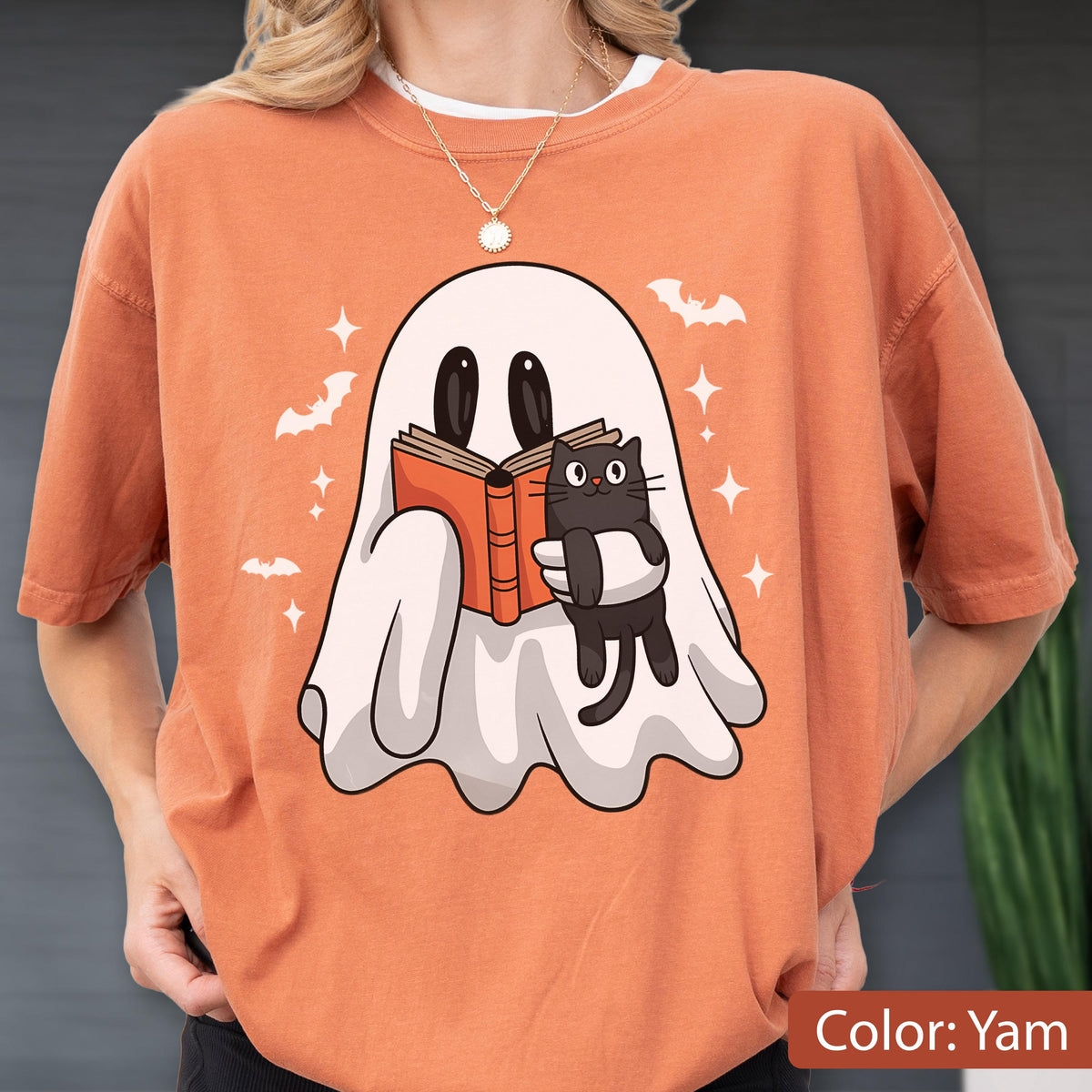 May include: Orange t-shirt featuring a cartoon ghost reading a book with a black cat. The ghost is white with black eyes, and the book is orange. Bats and stars are also printed on the shirt. The shirt's color is "Yam".