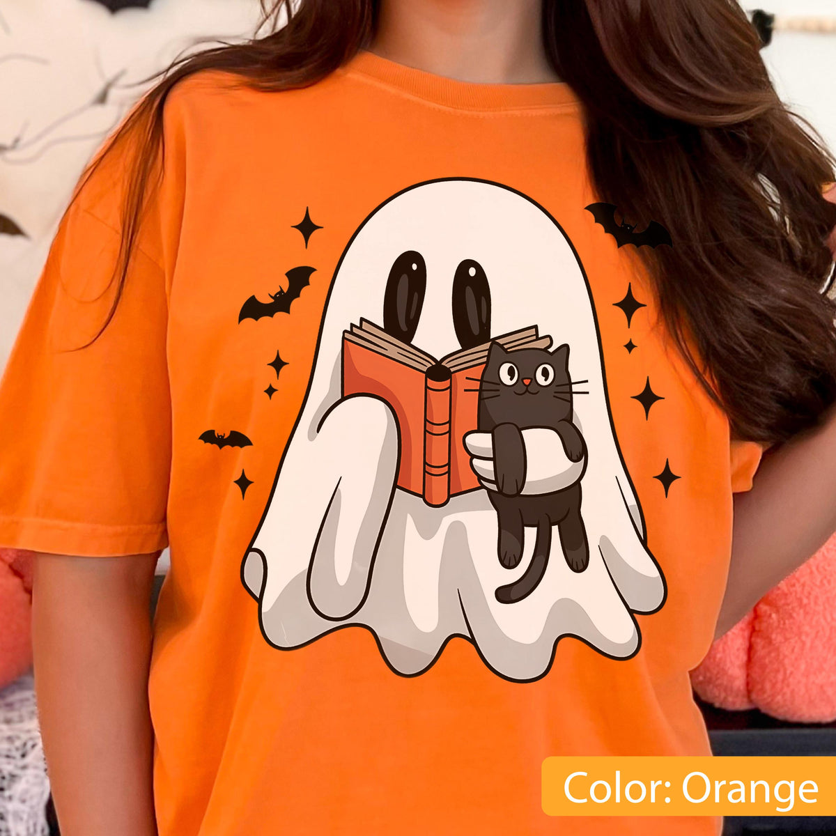 May include: Orange t-shirt featuring a cartoon ghost reading a book with a black cat. The ghost and cat are holding the book together. Bats and stars are also printed on the shirt. The text "Color: Orange" is at the bottom.