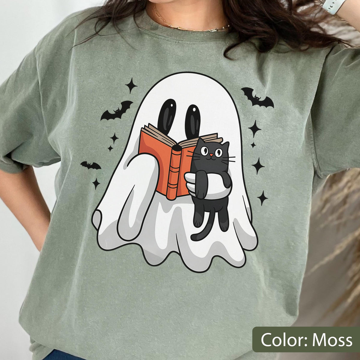 May include: Moss green t-shirt featuring a white ghost reading a book with a black cat. The ghost and cat are holding an orange book. Black bats and stars are scattered around the ghost. The text "Color: Moss" is at the bottom.