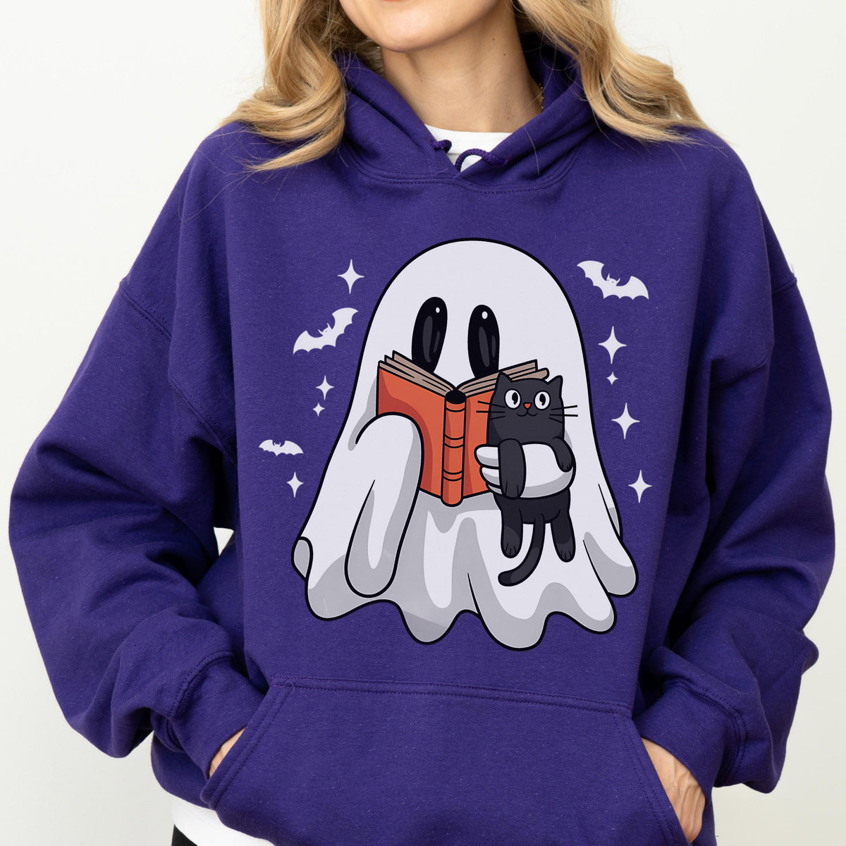 May include: A purple hoodie featuring a cartoon ghost reading a book and holding a black cat. The design includes bats and stars. The hoodie has a drawstring hood and a front pocket.