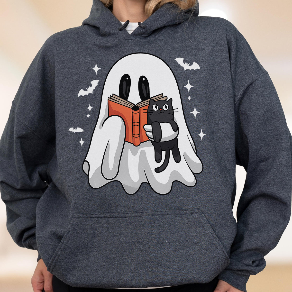May include: Dark gray hoodie featuring a cartoon ghost reading a book with a black cat. The ghost is white with black eyes and is holding an orange book. Bats and stars are also in the design. Halloween themed apparel.