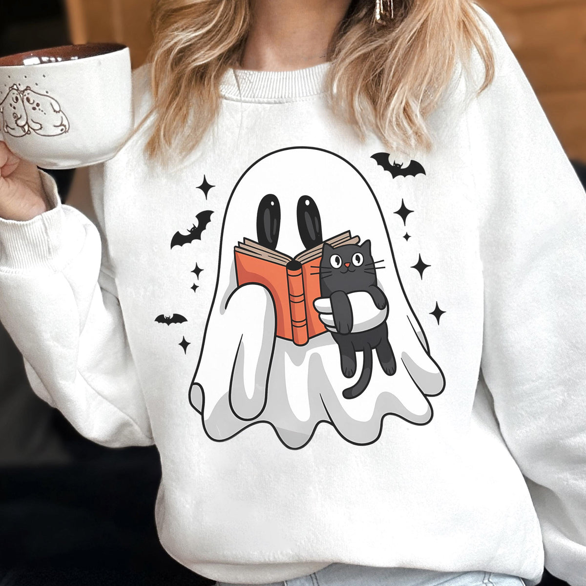 May include: White sweatshirt featuring a cartoon ghost reading a book with a black cat. The design includes bats and stars. The person is holding a white mug with a cartoon design.