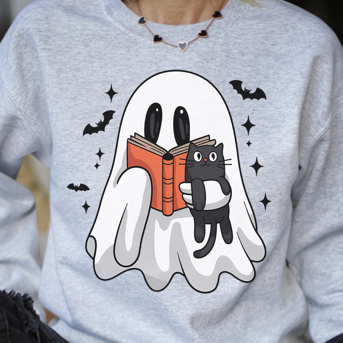 May include: A light gray sweatshirt featuring a white ghost reading a book with a black cat. The ghost and cat are illustrated in a cartoon style. Black bats and stars are scattered around the design.