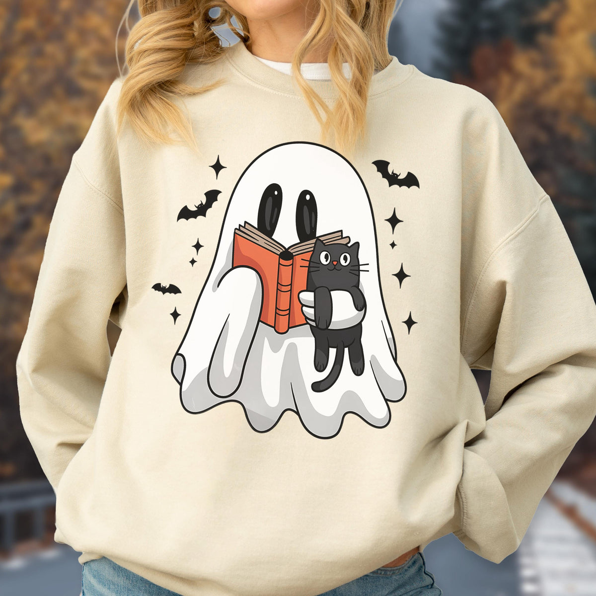 May include: Beige sweatshirt featuring a cartoon ghost reading a book with a black cat. The ghost is white with black eyes, holding an orange book. Black bats and stars surround the ghost, creating a Halloween theme.