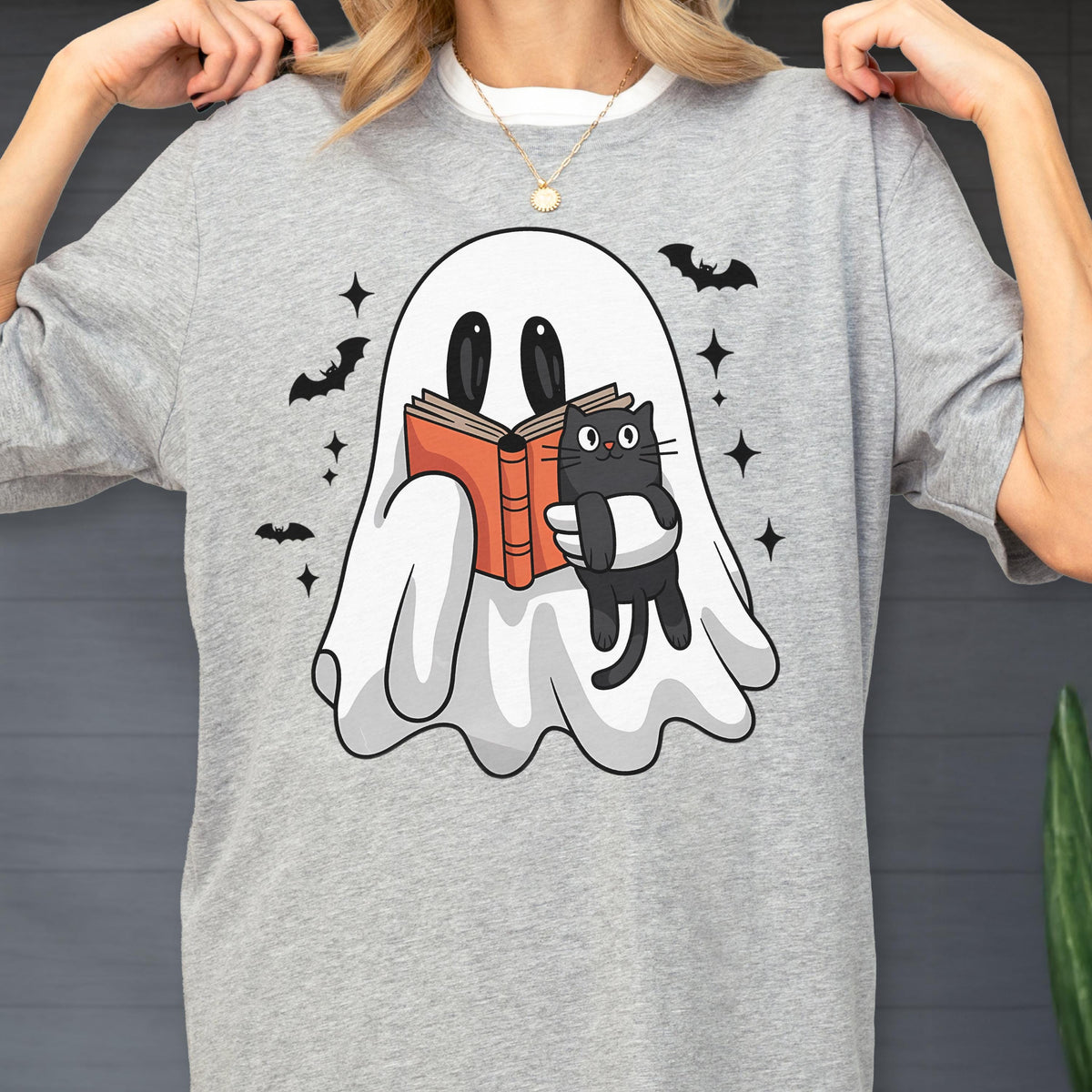 May include: A heather gray t-shirt featuring a graphic of a white ghost reading an orange book while holding a small black cat. The design is surrounded by black silhouettes of flying bats and small four-pointed star shapes scattered around the ghost.