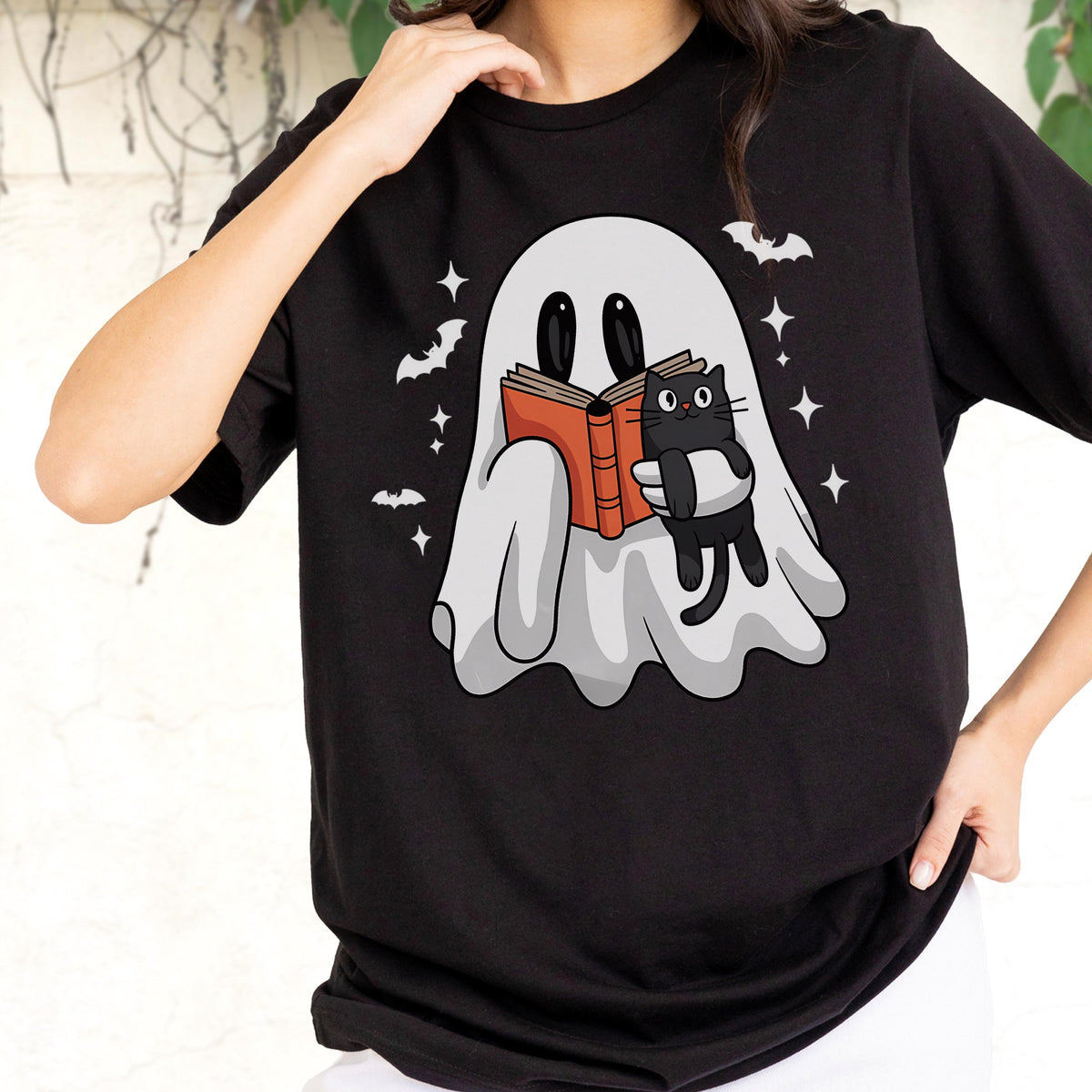 May include: A black t-shirt featuring a graphic of a white ghost reading an orange book while holding a small black cat. The design includes small white stars and bats surrounding the ghost, set against the solid black fabric of the shirt.