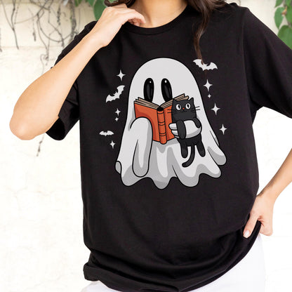 May include: A black t-shirt featuring a graphic of a white ghost reading an orange book while holding a small black cat. The design includes small white stars and bats surrounding the ghost, set against the solid black fabric of the shirt.