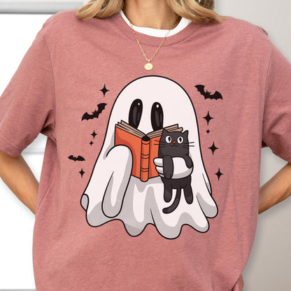 May include: A dusty rose t-shirt featuring a graphic of a white ghost reading an orange book while holding a black cat. The design is surrounded by small black bats and four-pointed stars. The shirt has a crew neckline and short sleeves.