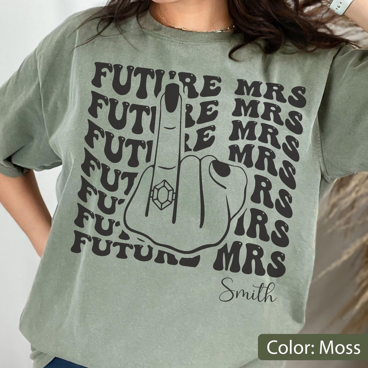 May include: A moss green t-shirt featuring a black graphic of a hand making a middle finger gesture with a diamond ring on the finger. The text "FUTURE MRS" is repeated in a wavy pattern behind the hand, with the name "Smith" printed below.