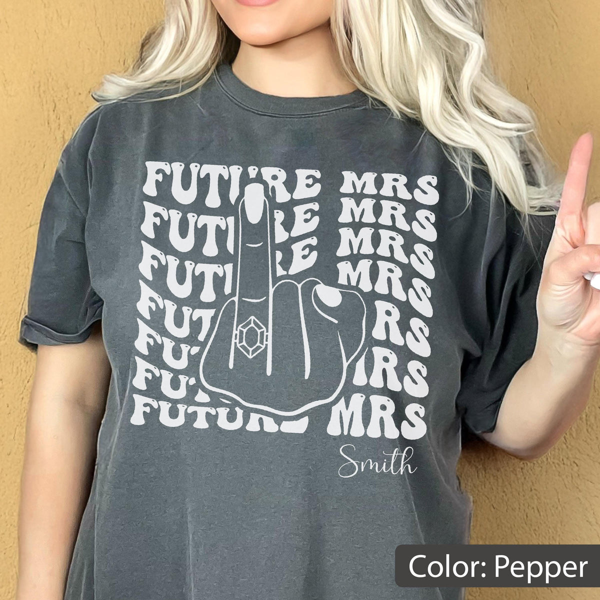 May include: A pepper-colored t-shirt featuring a white graphic of a hand making a middle finger gesture with a ring on the finger. The text "FUTURE MRS" is repeated in a wavy pattern behind the hand, and the name "Smith" is printed below.