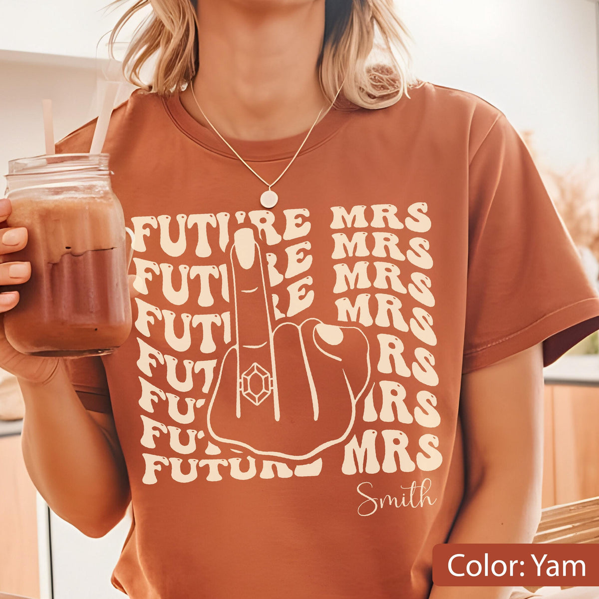 May include: A yam-colored t-shirt featuring a cream-colored graphic of a hand with a ring on the middle finger. The text reads FUTURE MRS repeatedly in a wavy pattern, with the name Smith printed in a cursive script at the bottom right corner.