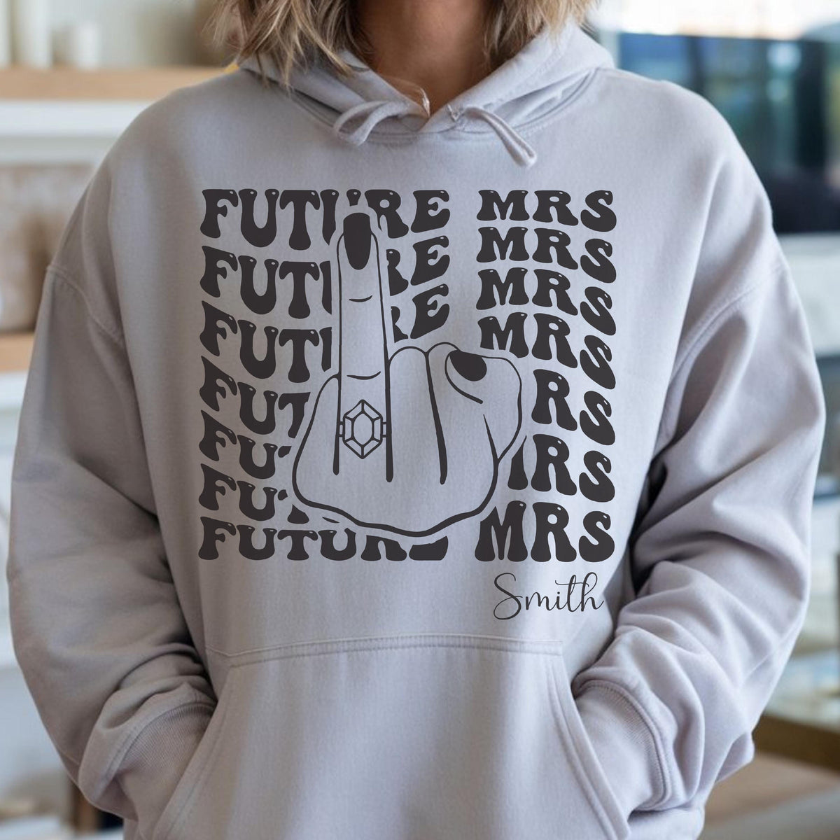 May include: A light gray hoodie with a front graphic. The graphic features the words "FUTURE MRS" and "MRS" in a wavy font, surrounding a hand with a ring on the middle finger. The name "Smith" is written below the hand.
