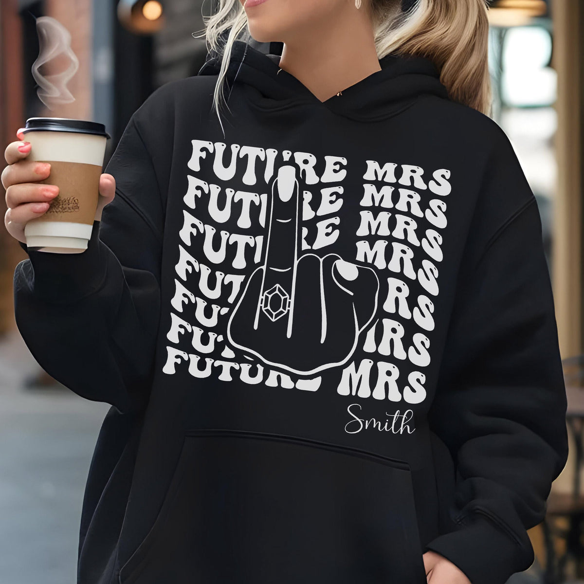 May include: Black hoodie with the text "FUTURE MRS" repeated in a wavy pattern. A graphic of a hand with a ring on the middle finger is in the center. The name "Smith" is below the graphic.