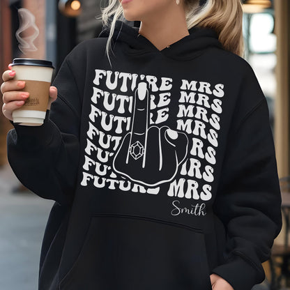 May include: Black hoodie with the text "FUTURE MRS" repeated in a wavy pattern. A graphic of a hand with a ring on the middle finger is in the center. The name "Smith" is below the graphic.