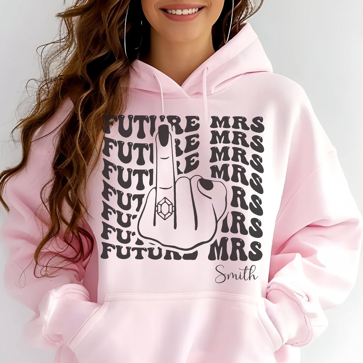 May include: A light pink hoodie with a graphic design. The design features the words "FUTURE MRS" and "MRS" in black, along with a hand giving the middle finger, wearing a diamond ring. The name "Smith" is printed below.
