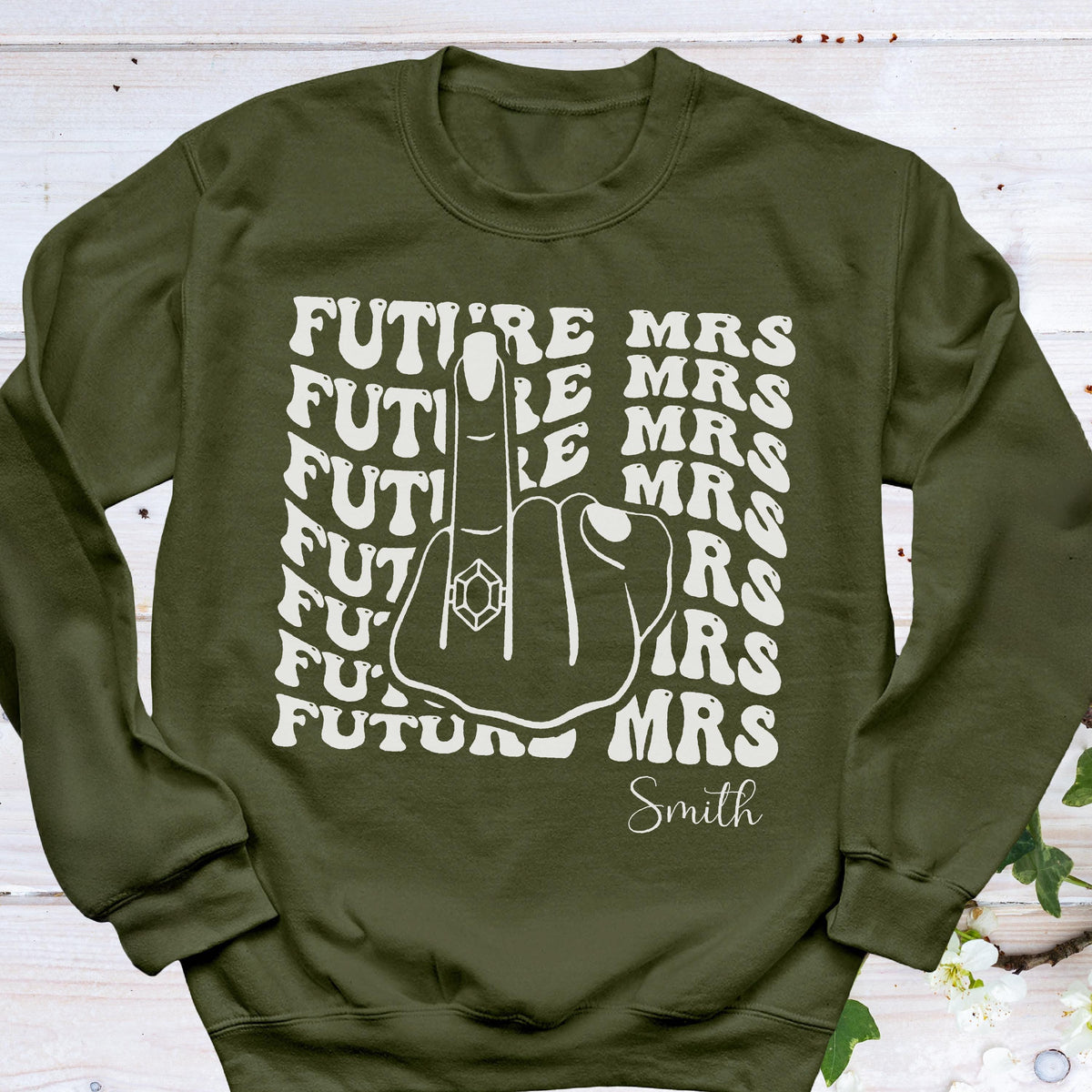 May include: Olive green sweatshirt with a white graphic. The graphic features a hand with a ring on the middle finger, surrounded by the words "FUTURE MRS" and the last name "Smith". The sweatshirt has a crew neck and long sleeves.