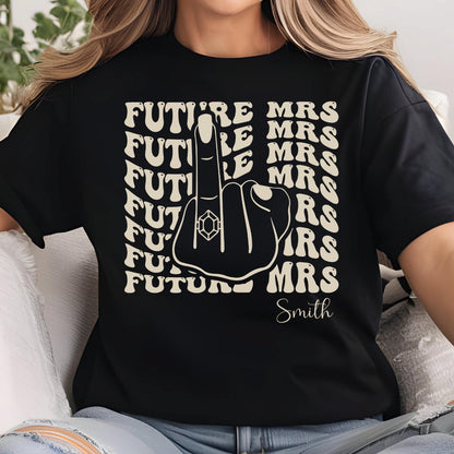 May include: A black t-shirt featuring a graphic of a hand making a middle finger gesture with a ring on the finger. The text "FUTURE MRS" is repeated in a wavy pattern in the background, and the name "Smith" is printed in script below the hand.