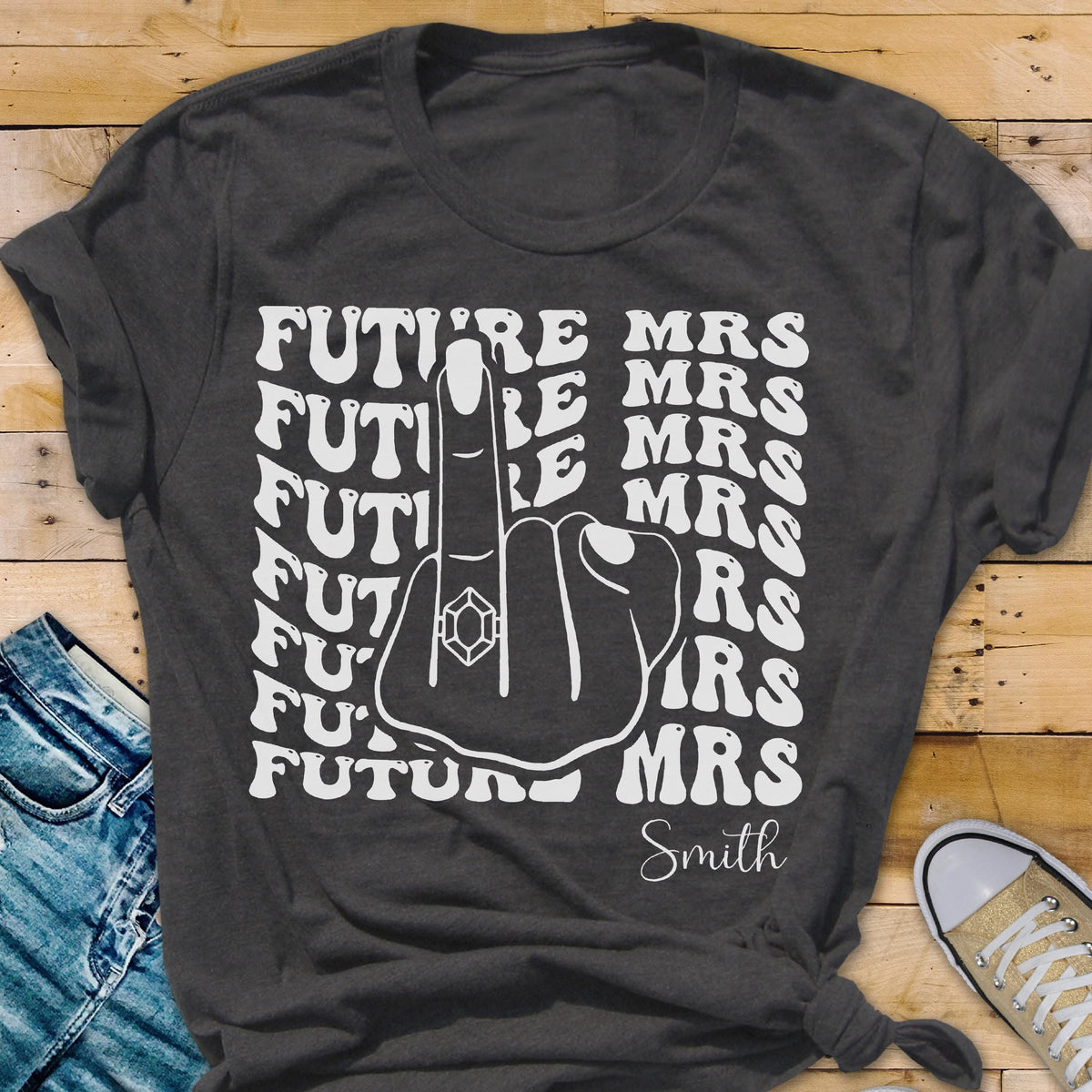 May include: A dark gray short-sleeve t-shirt featuring a white graphic of a hand with a ring on the middle finger. The text "FUTURE MRS" is repeated in a wavy pattern on both sides of the hand, with the name "Smith" printed below in script.