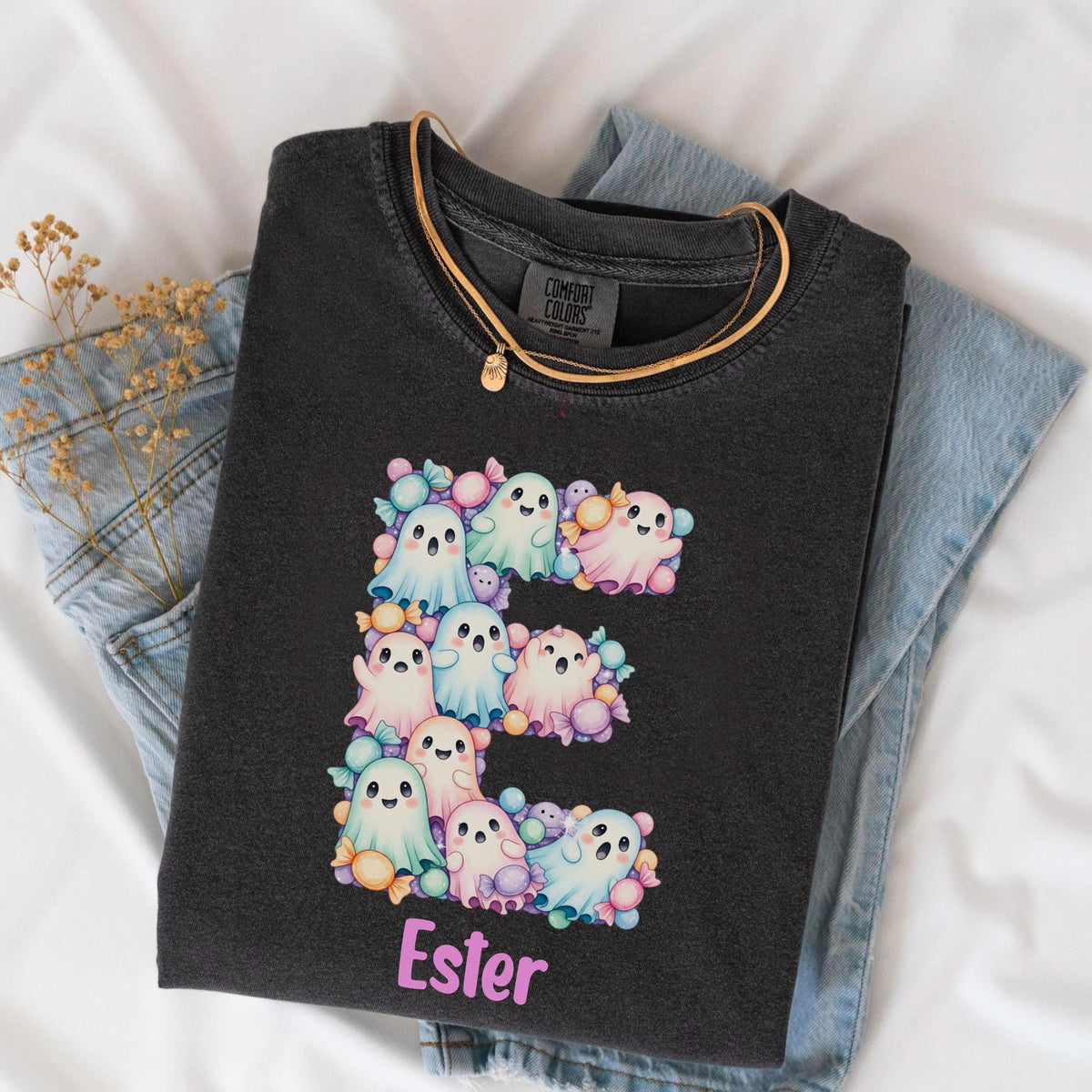 May include: A charcoal gray t-shirt featuring a large letter E composed of pastel-colored cartoon ghosts and wrapped candies. Below the letter, the name Ester is printed in light pink lettering. The shirt has a Comfort Colors brand label.