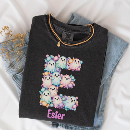 May include: A charcoal gray t-shirt featuring a large letter E composed of pastel-colored cartoon ghosts and wrapped candies. Below the letter, the name Ester is printed in light pink lettering. The shirt has a Comfort Colors brand label.