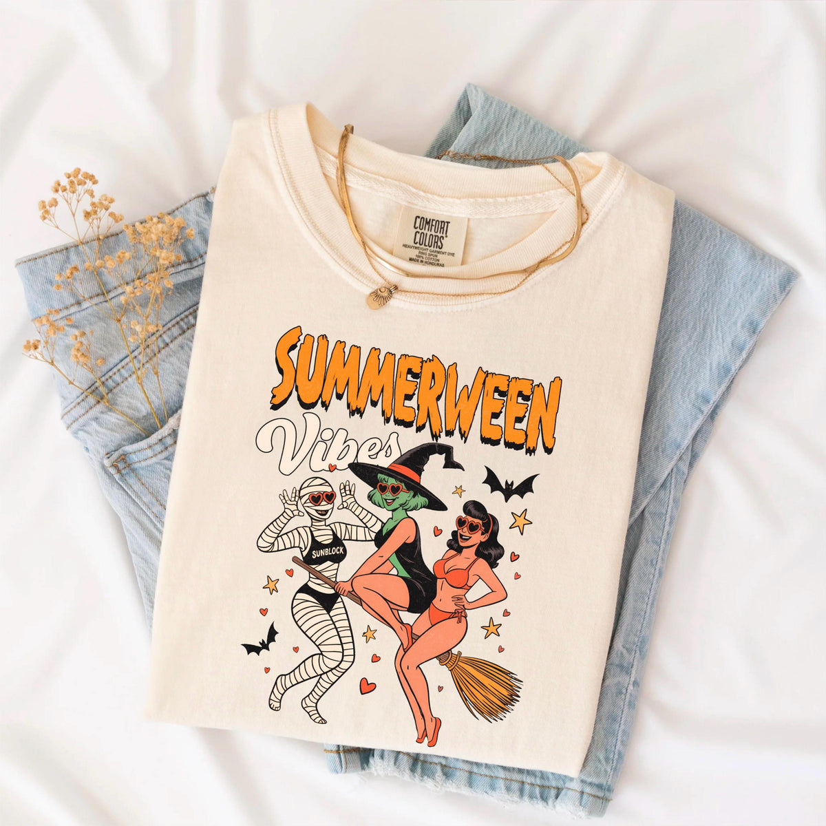 May include: Cream-colored t-shirt with the words "Summerween Vibes" in orange and white. The graphic features three cartoon characters: a mummy, a witch riding a broom, and a woman in a swimsuit. The shirt is folded on top of blue jeans.