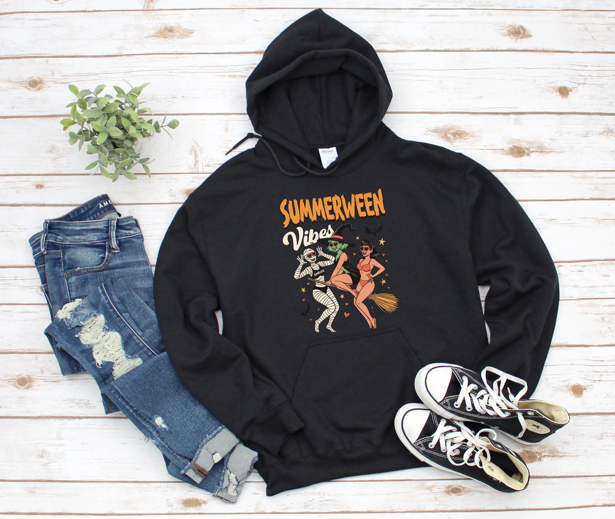 May include: Black hooded sweatshirt with the text "Summerween Vibes" and a graphic of three cartoon figures. The figures include a mummy, a witch, and a woman riding a broom. The sweatshirt is paired with blue jeans and black sneakers.
