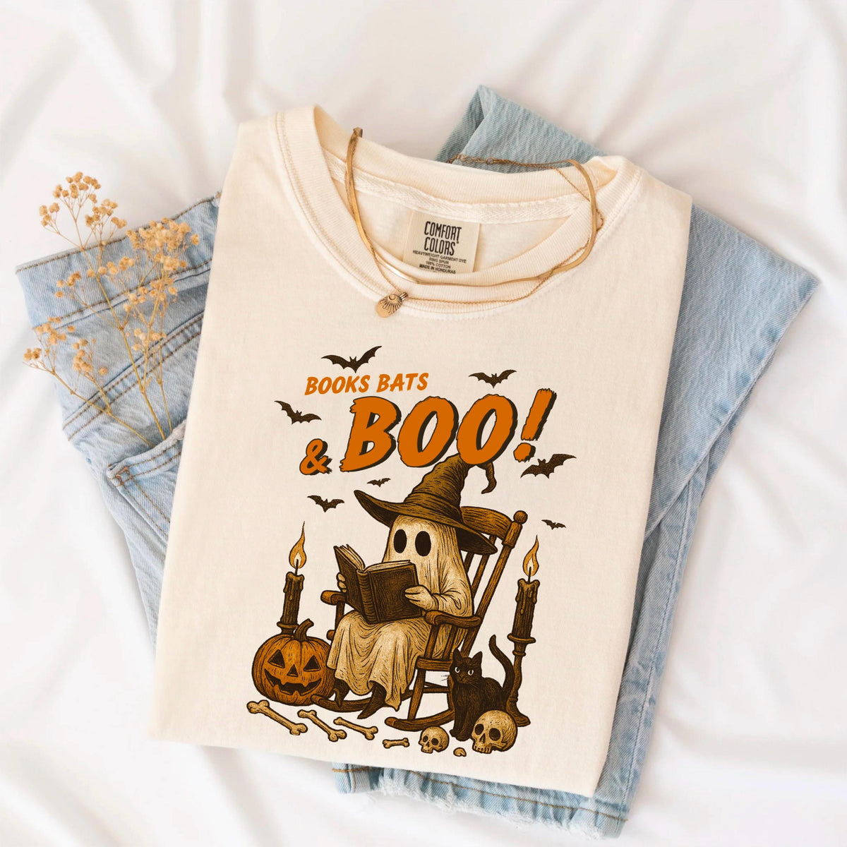 May include: Cream-colored t-shirt featuring a Halloween-themed graphic. The design includes a ghost reading a book, bats, a pumpkin, and the text "Books Bats & Boo!". The shirt is folded on top of blue jeans.