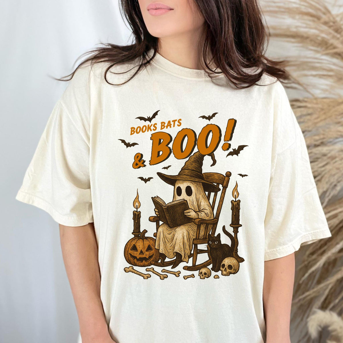 May include: Cream-colored t-shirt with a Halloween-themed graphic. The design features a ghost reading a book in a rocking chair, surrounded by bats, candles, a jack-o'-lantern, and the text "BOOKS BATS & BOO!".