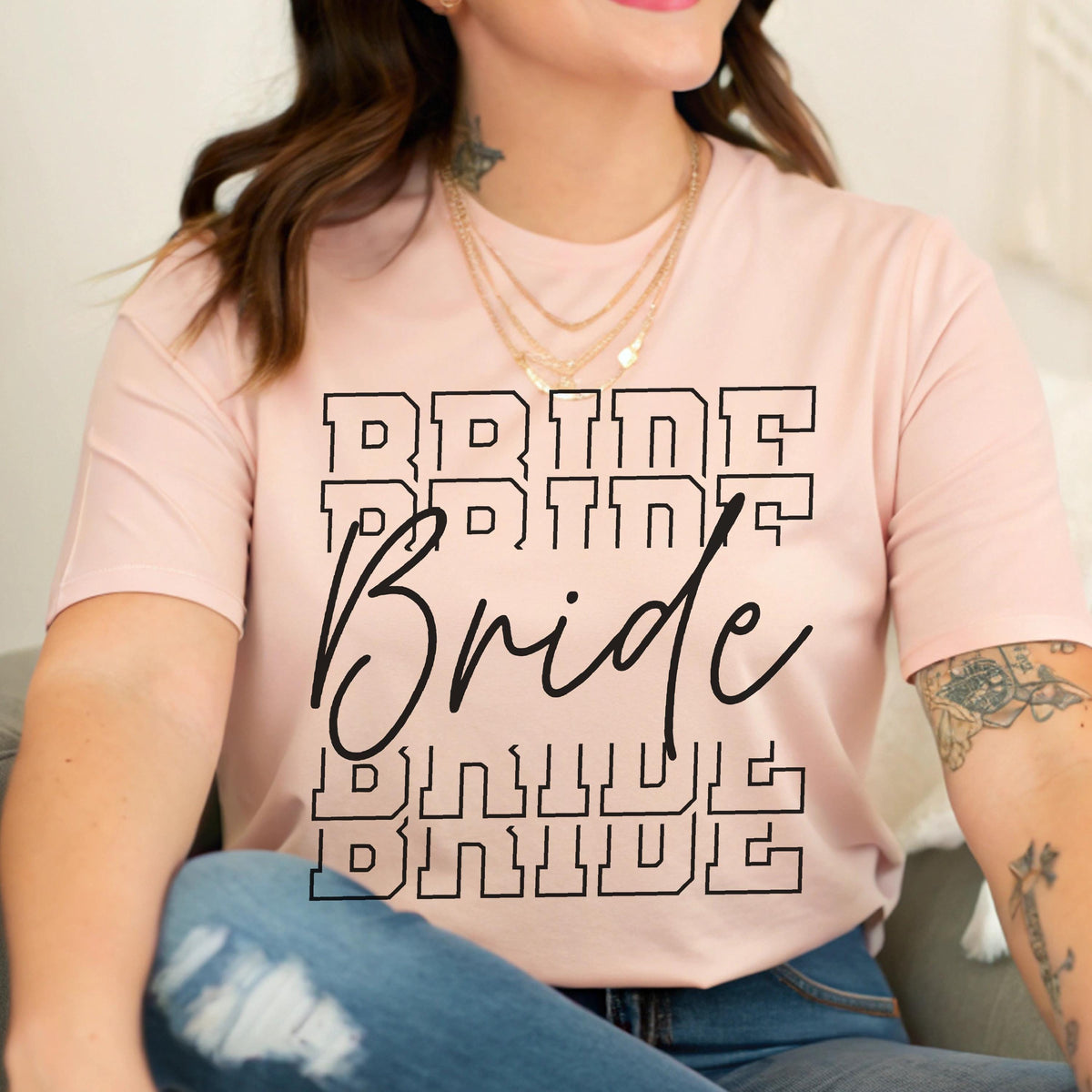 May include: A light pink t-shirt with the word "Bride" in a black, stacked, block letter design. The word "Bride" is also written in a cursive font. The shirt is a crew neck style.
