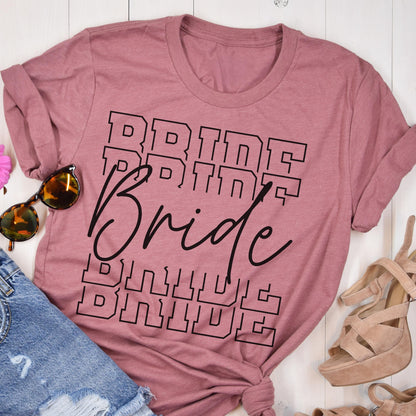 May include: A dusty rose t-shirt with the word "Bride" printed in black. The word is repeated in a stacked design, with a cursive "Bride" overlaying the stacked text. Sunglasses, denim shorts, and platform sandals are also visible.
