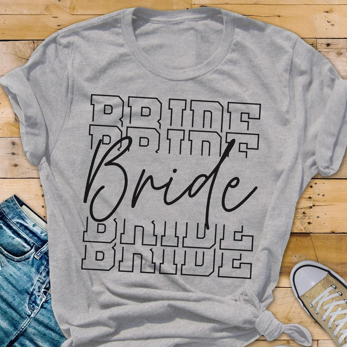 May include: A light gray t-shirt with the word "Bride" in a stacked design. The word "Bride" is in a cursive font, with the word "Bride" stacked in a block letter font above and below it. A pair of blue jeans and a sneaker are visible.