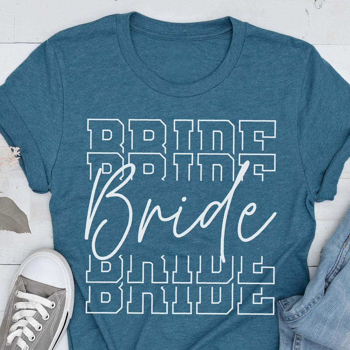 May include: Teal-colored t-shirt with the word "Bride" in white, in a stacked design. The word "Bride" is written in a cursive font, with the word "Bride" stacked above and below in a block letter font.